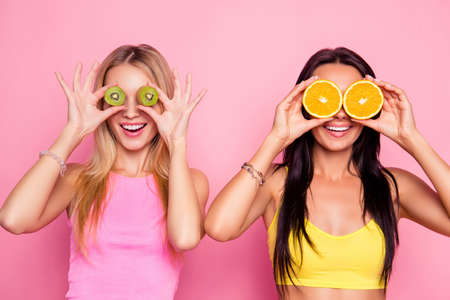 Surprised wondered astonished attractive pretty happy smiling charming joyful funky women are fooling around and covering their eyes with juice kiwi and orange, isolated on bright pink backgroundの写真素材
