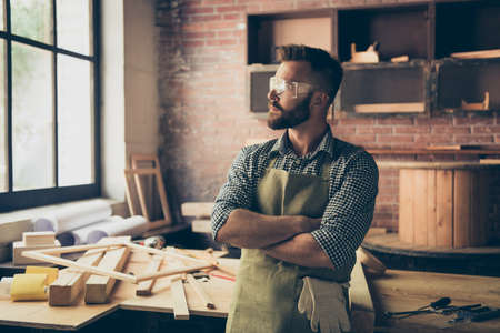 Portrait of pensive handsome bearded minded calm confident experienced carpenter standing in front of his work-table with folded arms, he is looking asideの写真素材
