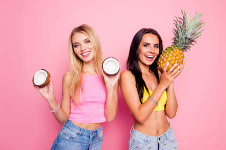 Slim attractive beautiful healthy excited smiling playful flirty sexy coquette girlfriends are holding exotic tropical fruit dressed in tight apparel and rejoicing, isolated on bright pink backgroundの写真素材