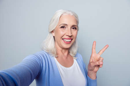 Close up portrait of charming happy cheerful with toothy beaming smile mature woman granny grandma grandmother demonstrating v-sing and making self portrait, isolated on grey background, copy-spaceの写真素材
