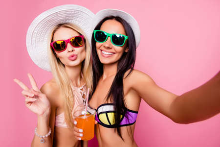 pretty tourists shooting selfie on front camera, showing two fingers with pout lips, having alcohol beverage, isolated on pink backgroundの写真素材