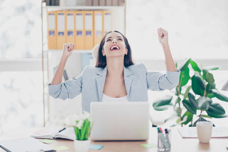 Pretty, laughing lawyer with raised arms and fists celebrating successfully completed work, sitting on chair at her desk in work placeの写真素材