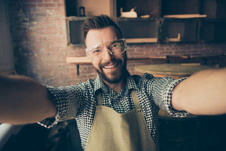 Close up portrait of happy cheerful joyful excited with toothy smile bearded wearing protective glasses carpenter, he is taking selfie in his workstationの写真素材