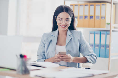 Pretty, charming, stylish, modern accountant in jacket using wi-fi, 3G internet on her smart phone, checking email, sitting in desktop in work stationの写真素材