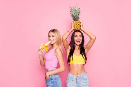 Happy beautiful smiling playful joyful cheerful women are eating banana and trying to hold a pineapple on a head, isolated on pink backgroundの写真素材