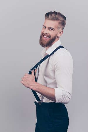 Vertical side profile half-turned portrait of cheerful excited careless classy rich confident laughing stylish careless guy correcting fixing his braces suspenders isolated on gray backgroundの写真素材