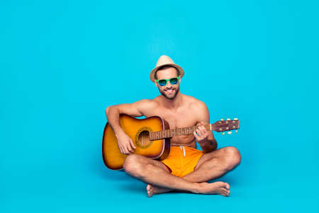Attractive, cheerful, talented guy with stubble in yellow shorts  sitting with crossed legs over blue background, having guitar in hands, playing songs, looking at camera, entertains the company of friendsの写真素材