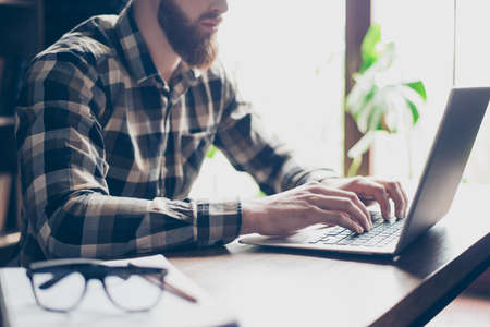 Cropped close up photo of serious concentrated handsome confident busy red bearded guy, hands fingers typing email on laptop sitting at the table in office near windowの写真素材