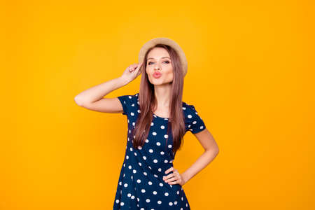 Portrait of sexy, nice, lovely, pretty, adorable woman in polka-dot dress holding hands on waist and hat, blowing air kiss at camera with pout lips, standing over yellow backgroundの写真素材