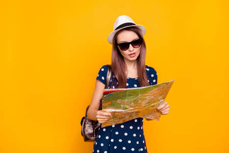 Portrait if thoughtful, pretty, charming woman in polka-dot dress having rucksack on shoulder, holding map in hand, trying to find a right way, standing over yellow backgroundの写真素材