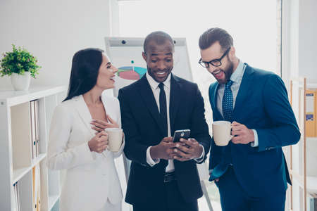 Stylish, modern, cheerful partners in tux, tuxedo having break time, watching comic video during coffee time on smart phone, standing in work place, station, holding mug with coffee in handsの写真素材
