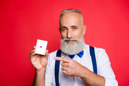 Professional, successful, handsome, attractive old man with cunning expression holding deck  in hand and pointing with forefinger on cards, looking at camera, isolated on red backgroundの写真素材