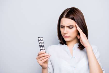 Pretty, charming, stylish, brunette, upset, sad, unhappy woman, touching her temple with hand, looking at effective plate of tablet, reading manual of medical product from diseases on grey backgroundの写真素材