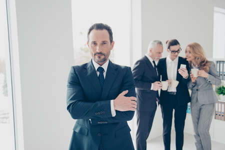 Portrait of attractive, smart brunet director in tux with tie standing in work place, station with crossed arms looking at camera, colleagues drinking coffee, talking, speaking on blurred backgroundの写真素材