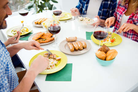 Top view cropped portrait of festive relatives imposing food in plates, sitting in house, apartment, room, having meat, sausages, bread, chops, salads and glasses of red wine on the tableの写真素材