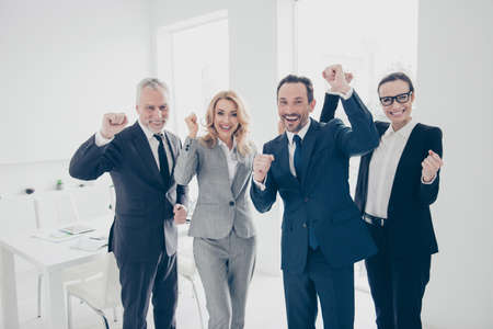 Portrait of attractive, lucky, stylish business people in suits celebrating victory in tender, successfully completed project with raised hands, looking at camera, standing on work place, stationの写真素材