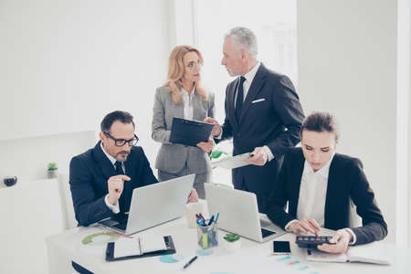 Portrait of concentrated, stylish, attractive business persons in suits working, sitting at desk, standing in work place, station, discussing, talking, speaking, using, laptop, clipboard, calculatorの写真素材