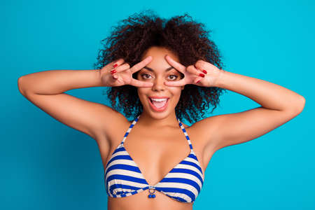 Close up portrait of cheerful mad careless slim fit with bronze skin afro american woman with curly hair showing double v-signs on her eyes, isolated on blue backgroundの写真素材