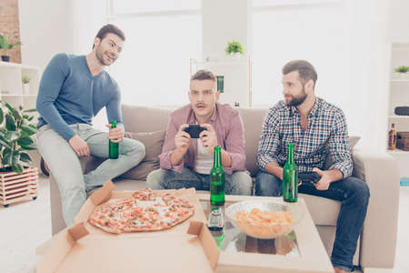 Three attractive handsome stylish men in casual outfits playing videogame, laughing from frustrated upset loser sitting in living room, drinking  beer, lager from bottle, having pizza, chipsの写真素材