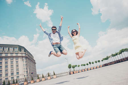 Bottom view low angle of funny crazy cheerful stylish trendy couple in spectacles jumping in the air showing two fingers, peace symbols, hands up, shouting screaming over sky clouds outdoor backgroundの写真素材