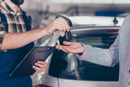 Close up cropped photo of professional smart experienced wearing uniform mechanic giving keys back to the owner with license of the fixed car, gray silver automobile is on the blurred background