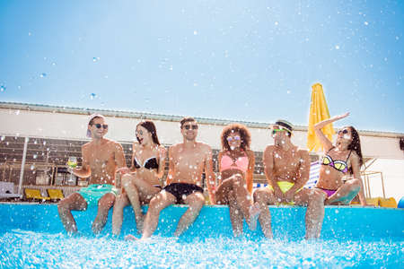 Party all day! Go crazy non stop. Six excited multi national tourists are enjoying at the pool sunny beach disco, with drinks, splashes of water all around, sitting by the pool, legs in clean waterの写真素材
