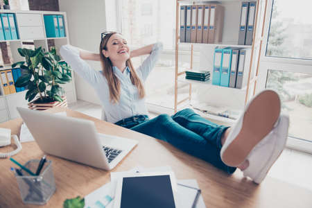 Portrait of attractive, stylish, cheerful, pretty woman holding hand behind the head, put legs on the table, having closed eyes, dreaming about vacation, weekend, rest, wearing pantsの写真素材