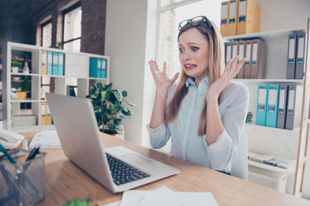 Portrait of charming, emotional, worried, nervous woman with glasses on head and hairstyle gesture with hands, looking at computer, something went wrong, can't get in time, sitting at deskの写真素材
