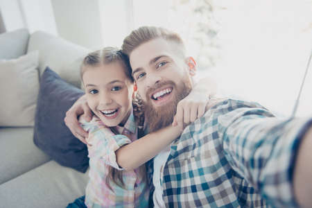 Self portrait of cheerful joyful family with one parent, laughing girl embracing her father with ginger hair shooting selfie on front camera sitting on couch in living room, enjoying time togetherの写真素材