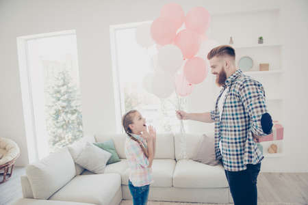 Stylish handsome father prepare gift case in pink package holding behind back  giving air balloons to wondered shocked amazed daughter together standing in modern white room with interiorの写真素材