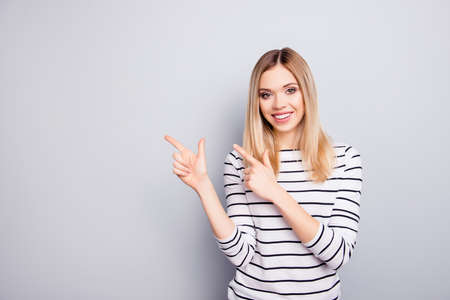 Portrait with empty place for advertisement of pretty, charming, cheerful, trendy, cute woman in striped outfit pointing two forefingers to copy space, looking at camera, isolated on grey backgroundの写真素材