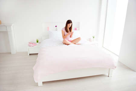 Attractive, pretty, cheerful, charming girl sit on bed with crossed legs having smart phone in hands, chatting with lover, texting sms, checking email, using 5G, wifi internet in open space bedroomの写真素材