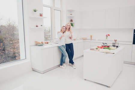 Full size portrait of mother and daughter standing in modern kitchen holding mugs with tea in hands while preparing salad vegetarian meal dinner having vegetables kitchenware on the tableの写真素材