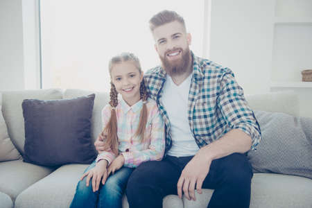 Portrait of cheerful positive family with one child parent, cute little girl with pigtails hairstyle attractive father with ginger hair in shirt sitting indoor in living room looking at cameraの写真素材