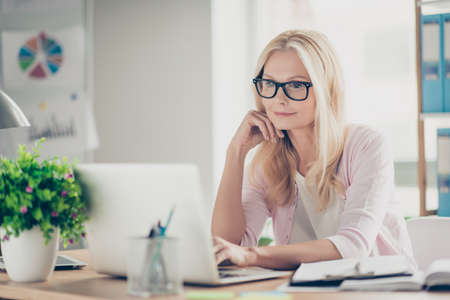 Nice, cute, confident, charming middle aged headmistress sitting at desk in workplace, holding hand near face, searching information on computer, checking email