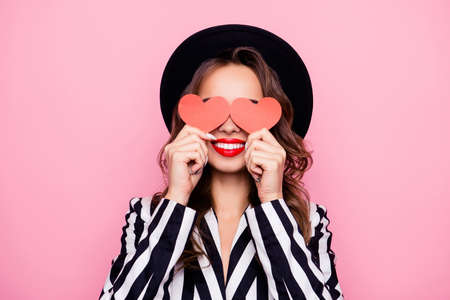 Charming pretty nice, comic, funny, modern, cheerful girl in black and white outfit having two small paper heart in hands on eyes place, isolated on pink backgroundの写真素材