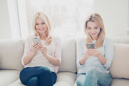 Portrait of cheerful positive mother and daughter sitting on couch in living room having smart phones in hands texting sms using 3G wi-fi internet searching contact technology apps conceptの写真素材