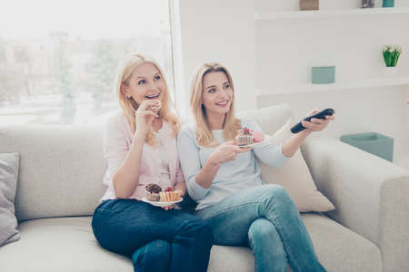 Portrait of cheerful attractive positive mother and daughter having cookies cakes muffins on saucer in hands searching film favorite program enjoying free time holiday using consoleの写真素材