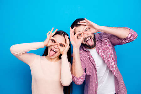 Portrait of cheerful joyful couple in casual outfit making binoculars with fingers gesturing tongue...