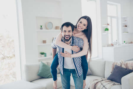 Portrait of playful foolish couple in casual outfits bearded man carrying his lover on back looking at camera in modern white apartment with interiorの写真素材