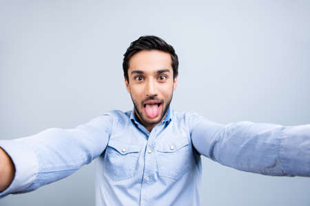 Self portrait of funny comic guy with black hair gesturing tongue out shooting selfie with two hands on front camera isolated on grey backgroundの写真素材