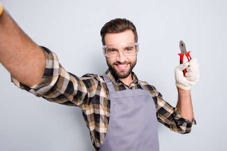 Self portrait of joyful cheerful man with stubble shooting selfie on front camera, showing pliers in arm, having fun, leisure, video-call, isolated on grey backgroundの写真素材