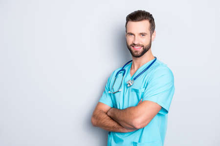 Portrait with copy space, empty place of positive cheerful man with stethoscope on his neck in blue lab uniform, having his arms crossed, isolated on grey backgroundの写真素材
