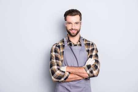 Portrait of attractive joyful carpenter with hairstyle in safety glasses looking at camera having his arms crossed standing over grey backgroundの写真素材