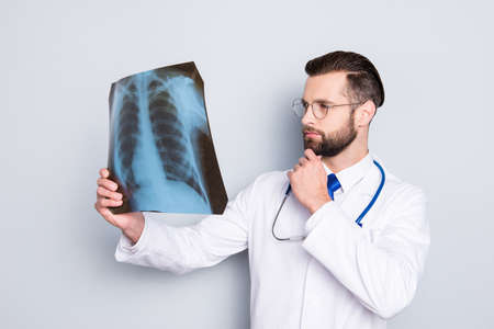 Portrait of pensive minded doc in white lab coat, uniform holding hand on chin looking at x-ray of...
