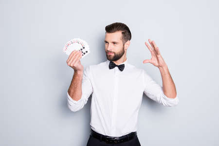 Portrait of cunning foxy magician with modern hairstyle and stubble in white shirt with bowtie holding set of cards in hand, making trick, focus, isolated on grey backgroundの写真素材