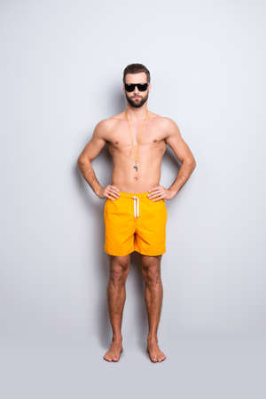 Full size body portrait of muscular athletic lifeguard with stubble in yellow shortsisolated on grey background, holding hands on waistの写真素材