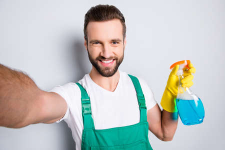 Self portrait of cheerful joyful cleaner with stubble in uniform shooting selfie on front camera having, showing detergent spray with blue liquid, isolated on grey backgroundの写真素材