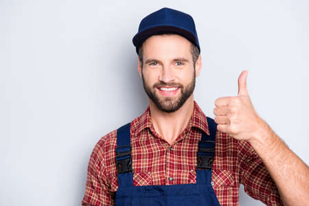 Close up portrait of handsome confident mechanic with stubble in shirt, overall, showing thumbup recommend approve advice sign, isolated on grey backgroundの写真素材