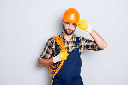 Portrait of virile harsh electrician with bristle in overall, shirt, having rolled cable on shoulder, holding hand on hardhat, standing over grey backgroundの写真素材